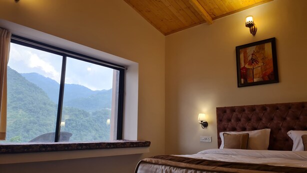 A hotel room with a large window offering a mountain view, a tufted headboard, a wall-mounted lamp, and a framed painting. - Prashanti Farms & Resort (Dehradun)