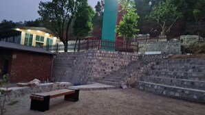 A courtyard with a stone wall, a wooden bench, and a metal fence. - Prashanti Farms & Resort (Dehradun)