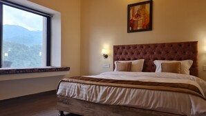 A bedroom with a large bed, a window with a view of mountains, a wall-mounted lamp, and a painting. - Prashanti Farms & Resort (Dehradun)