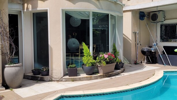 Una piscina con superficie de agua azul cristalina, rodeada por una plataforma de concreto. Se observan plantas en macetas y una parrilla de barbacoa al fondo.