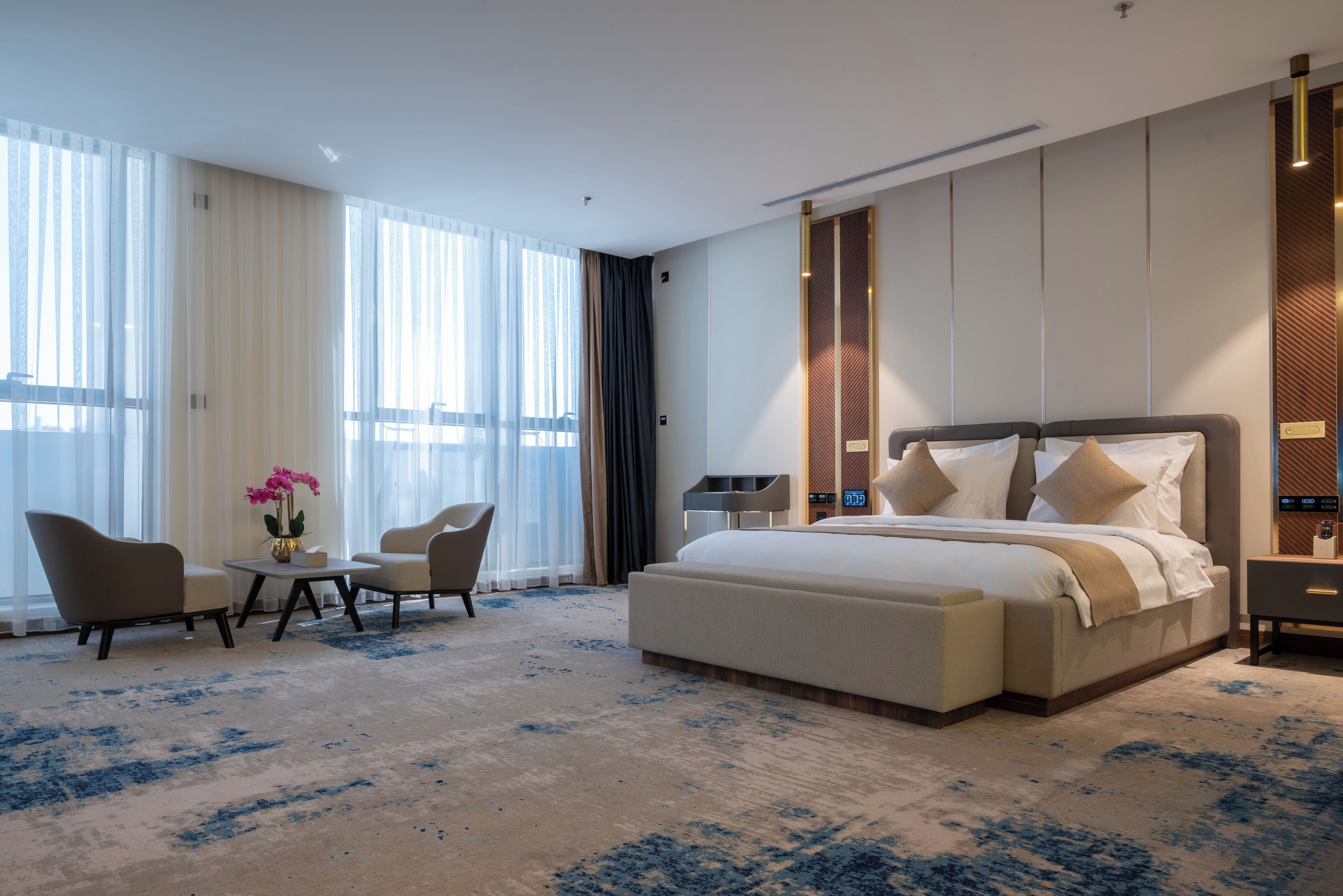 Une chambre d’hôtel moderne avec un grand lit, deux fauteuils, une petite table ornée d’un vase de fleurs et une grande fenêtre avec des rideaux légers.