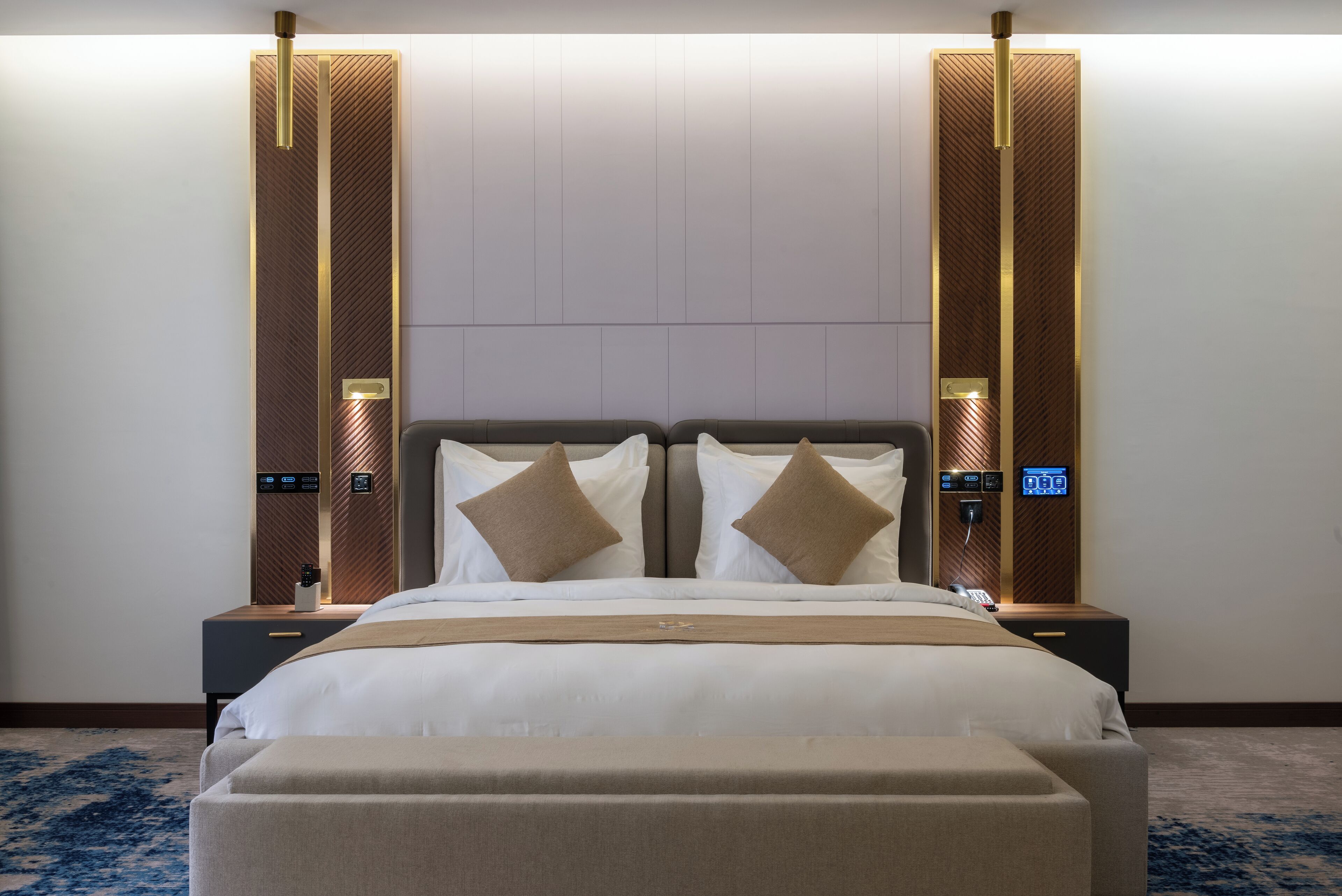 A modern hotel room with a large bed, two bedside tables, a digital clock, and a wall-mounted panel.