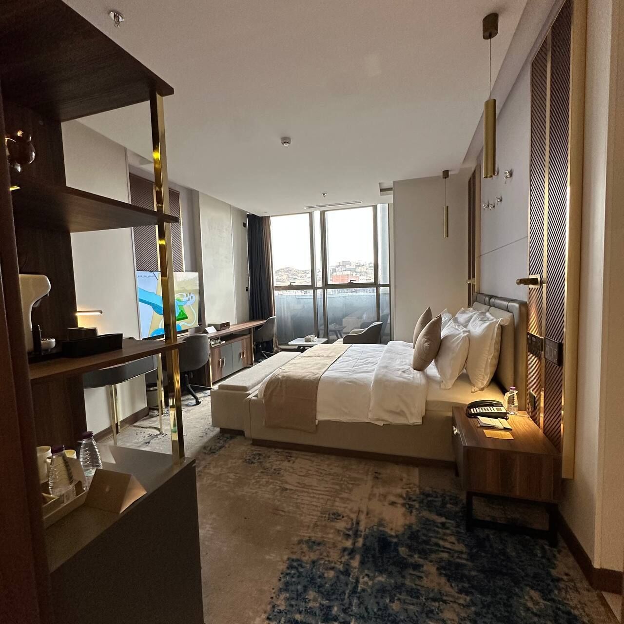 A modern hotel room with a large bed, a desk, a chair, and a view of the city through large windows.