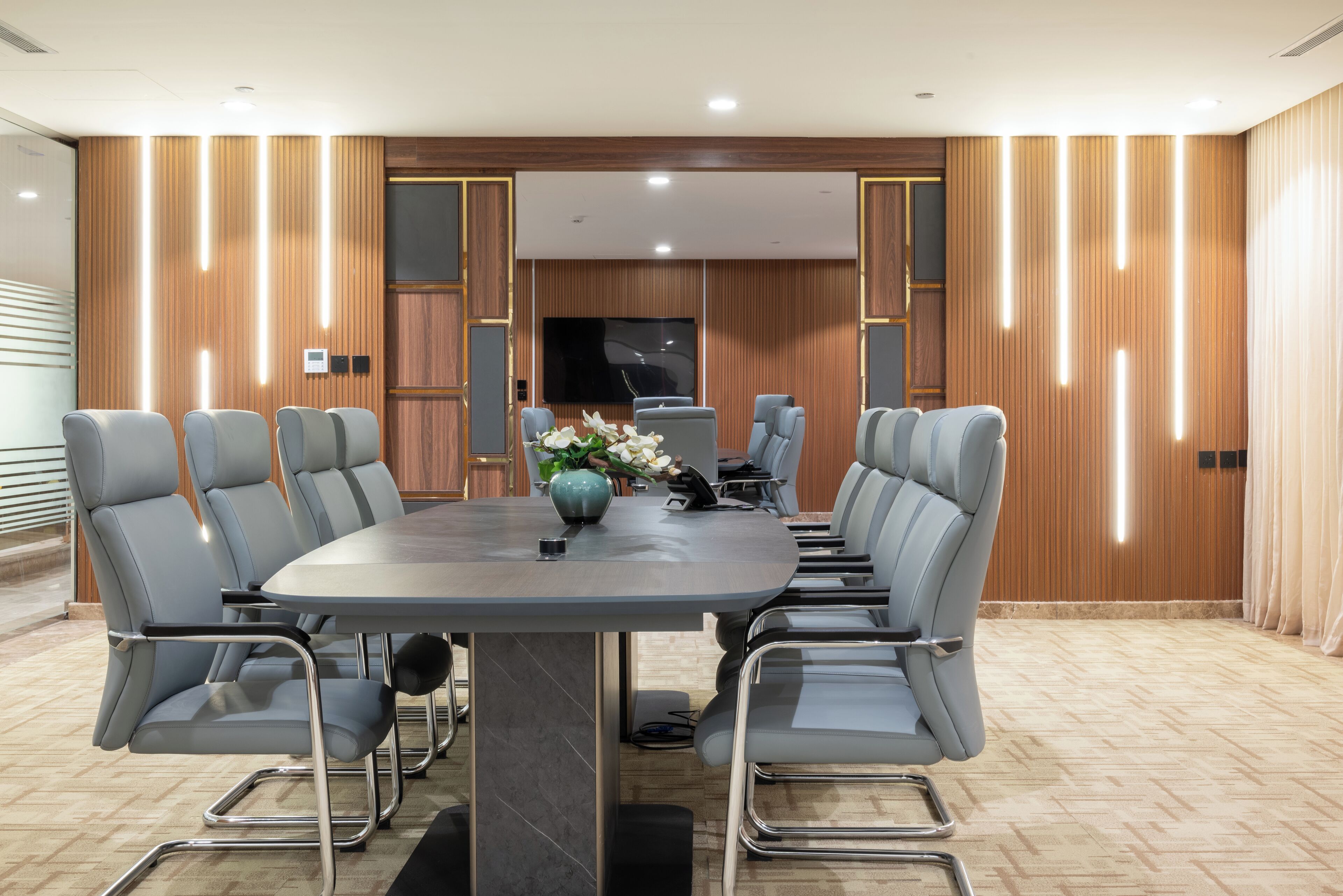 A conference room with a round table, grey chairs, a large flat-screen TV, and wooden paneling.