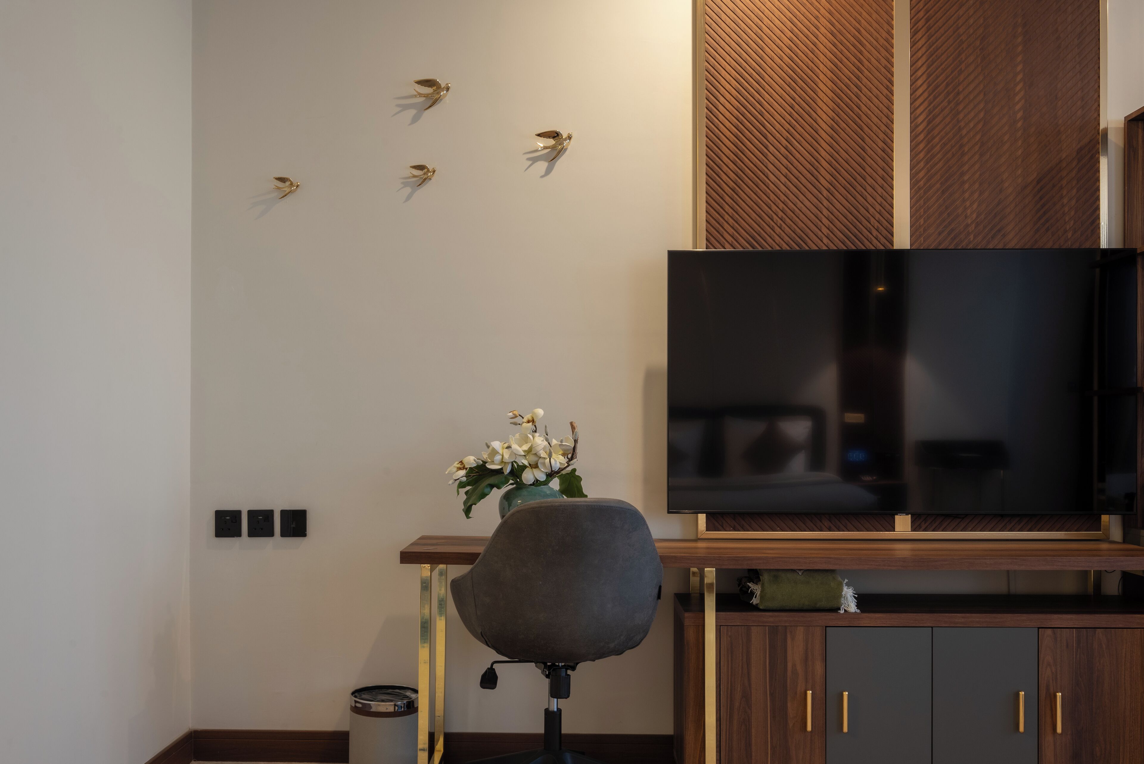 A modern living room with a flat-screen TV, a grey office chair, a wooden cabinet, and decorative wall art.