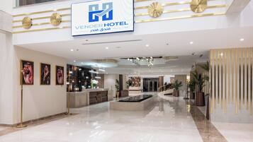 Un moderno vestíbulo de hotel con mostrador de recepción, obras de arte enmarcadas, fuente central y un cartel con el nombre del hotel.