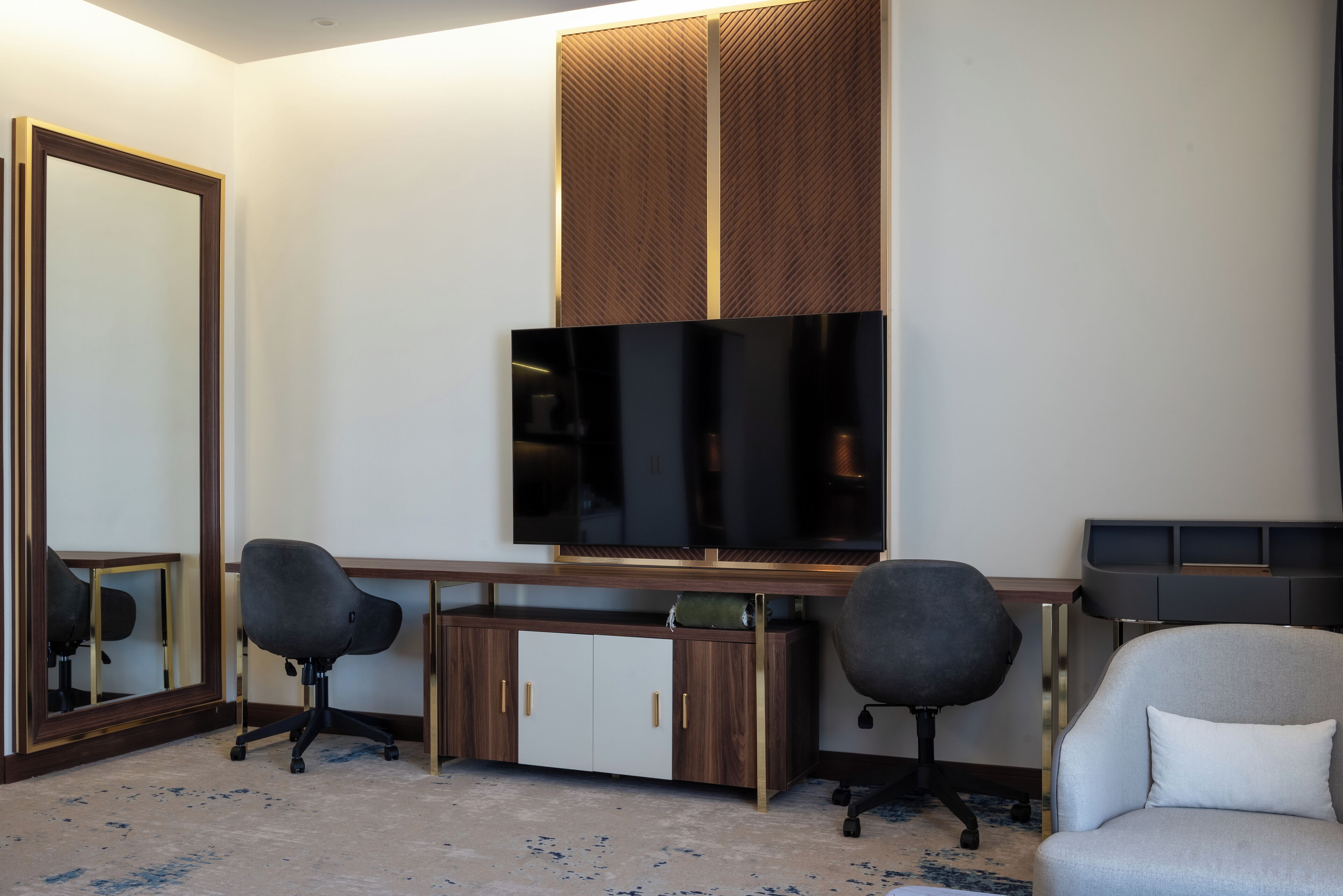 A hotel room with a large mirror, a flat-screen TV, a wooden desk, two office chairs, and a sofa.