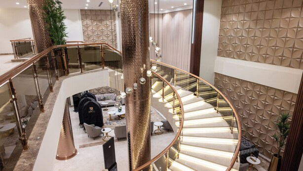 Un interno lussuoso con una scala a forma di ferro di cavallo, una zona salotto con divani e tavolini, e una parete decorata con motivi geometrici.