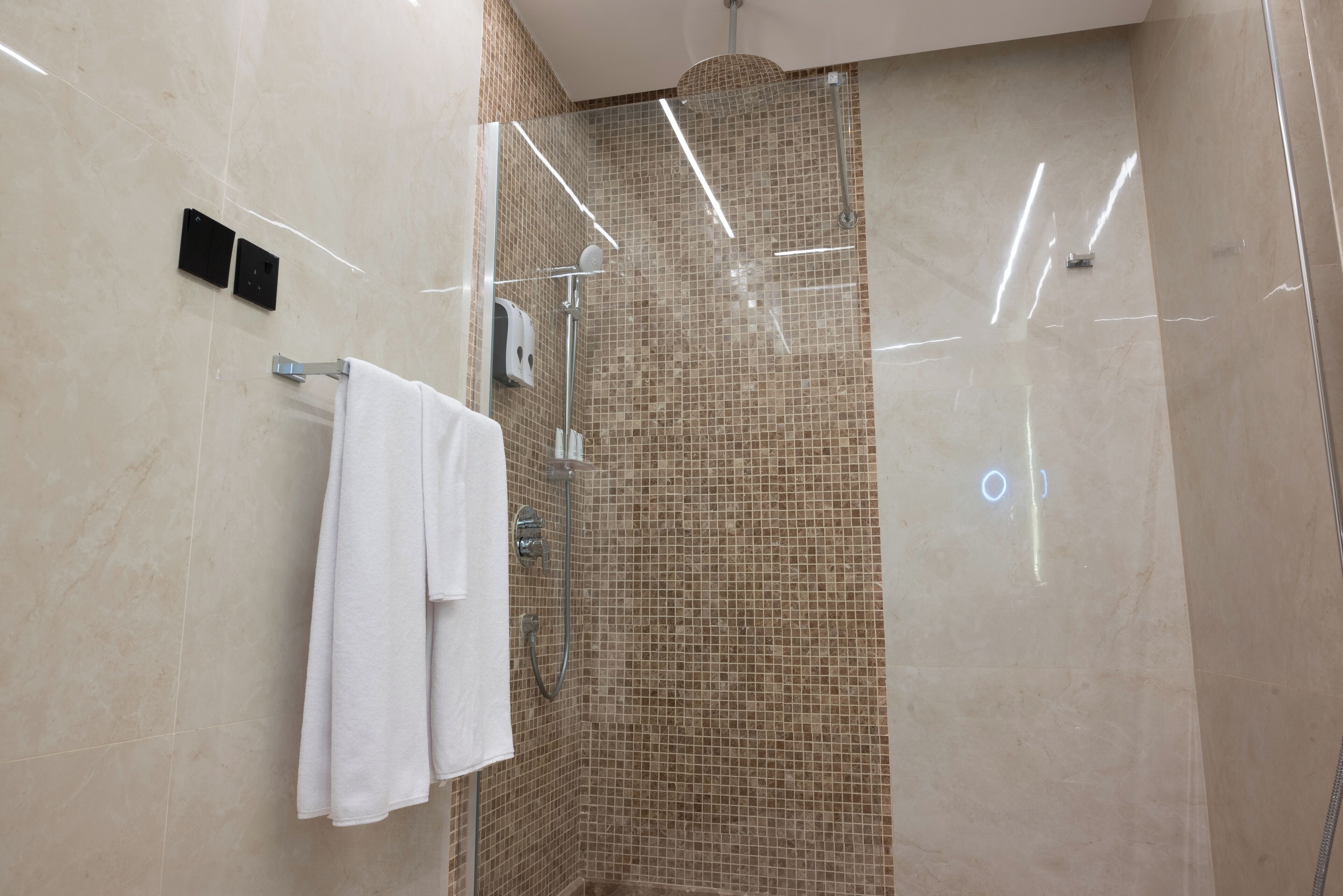 A modern bathroom with a glass-enclosed shower, a towel rack with two white towels, and a wall-mounted soap dispenser.
