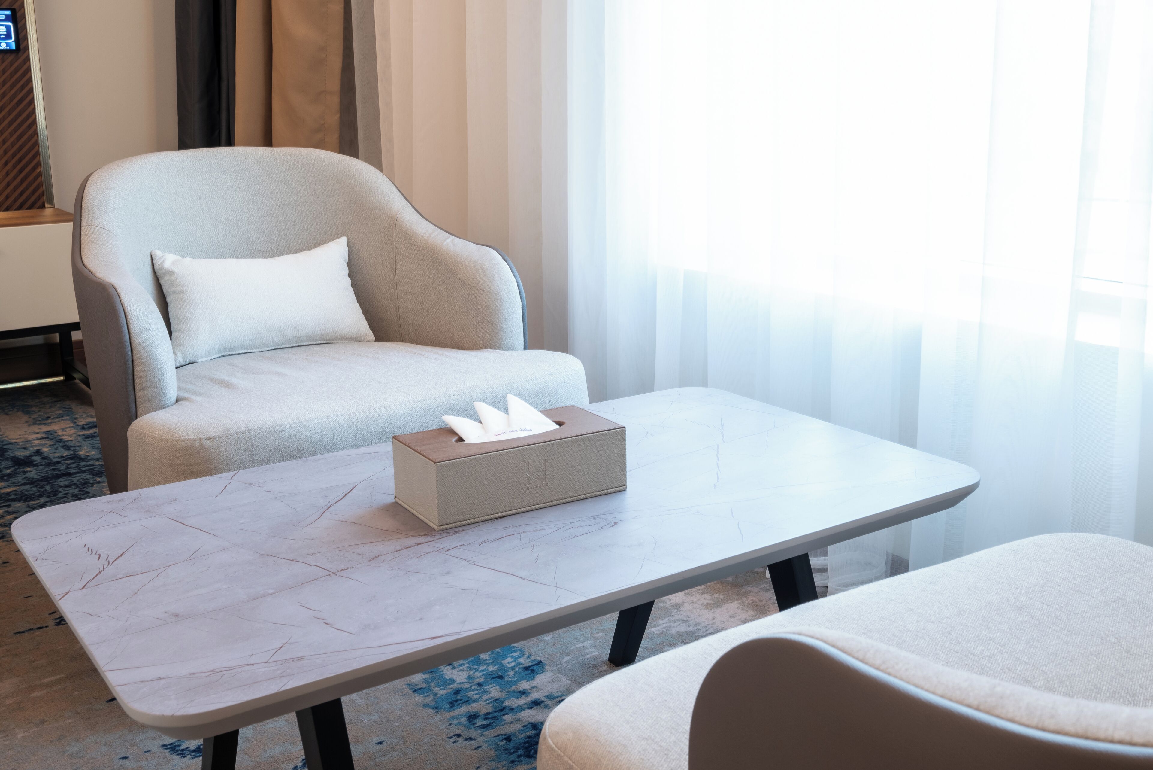 A modern hotel room with a light-colored armchair, a matching ottoman, a small table with a tissue box, and a large window with sheer curtains.