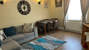 A cozy living room with a grey sofa, a patterned rug, a wall clock, a dining table with chairs, and a window with curtains. - Llanidloes - Sleeps 7 - Parking - Pet Friendly (Llanidloes)