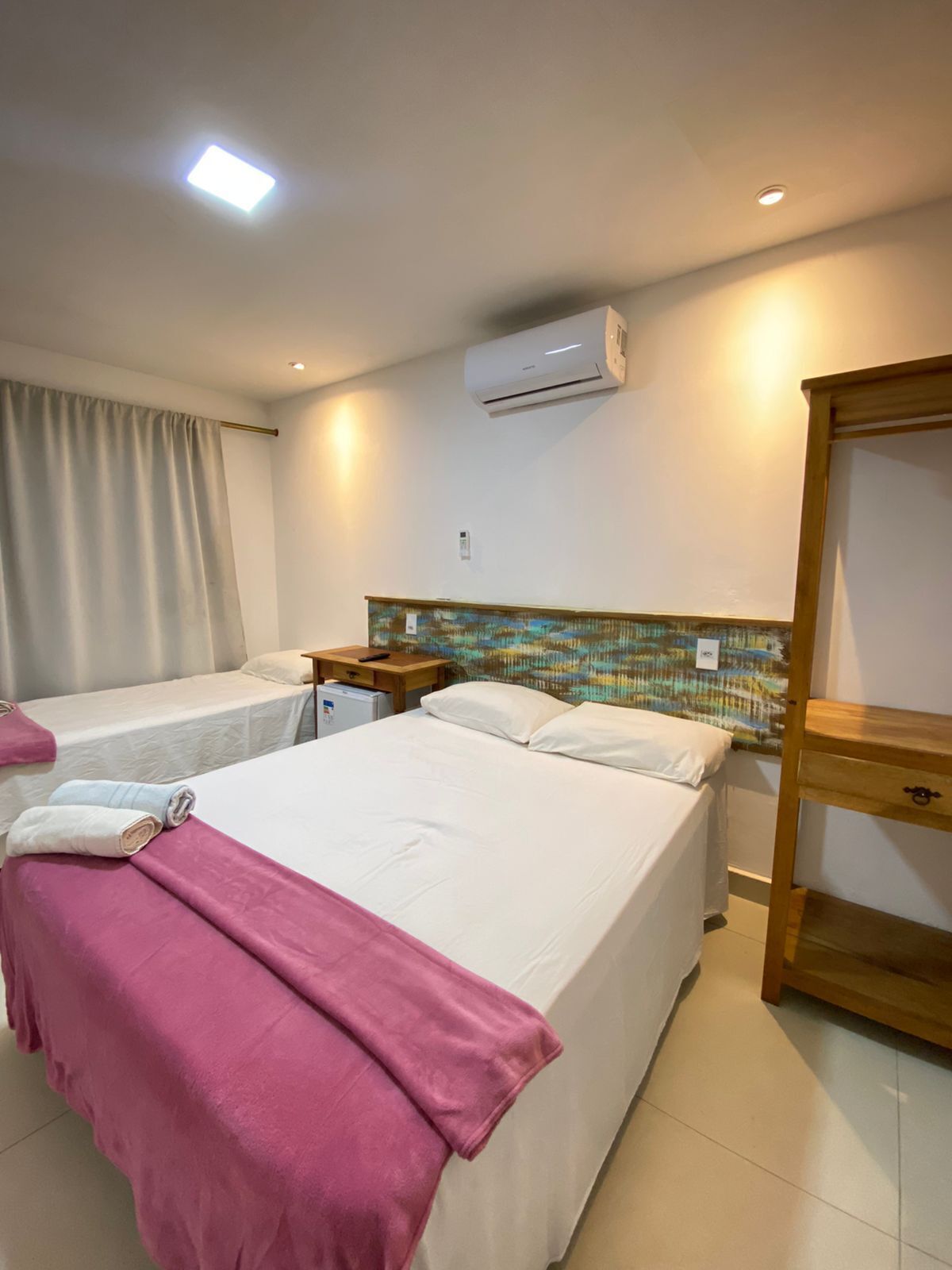 A hotel room with a large bed, a smaller bed, a wooden nightstand, an air conditioning unit, and a mirror.