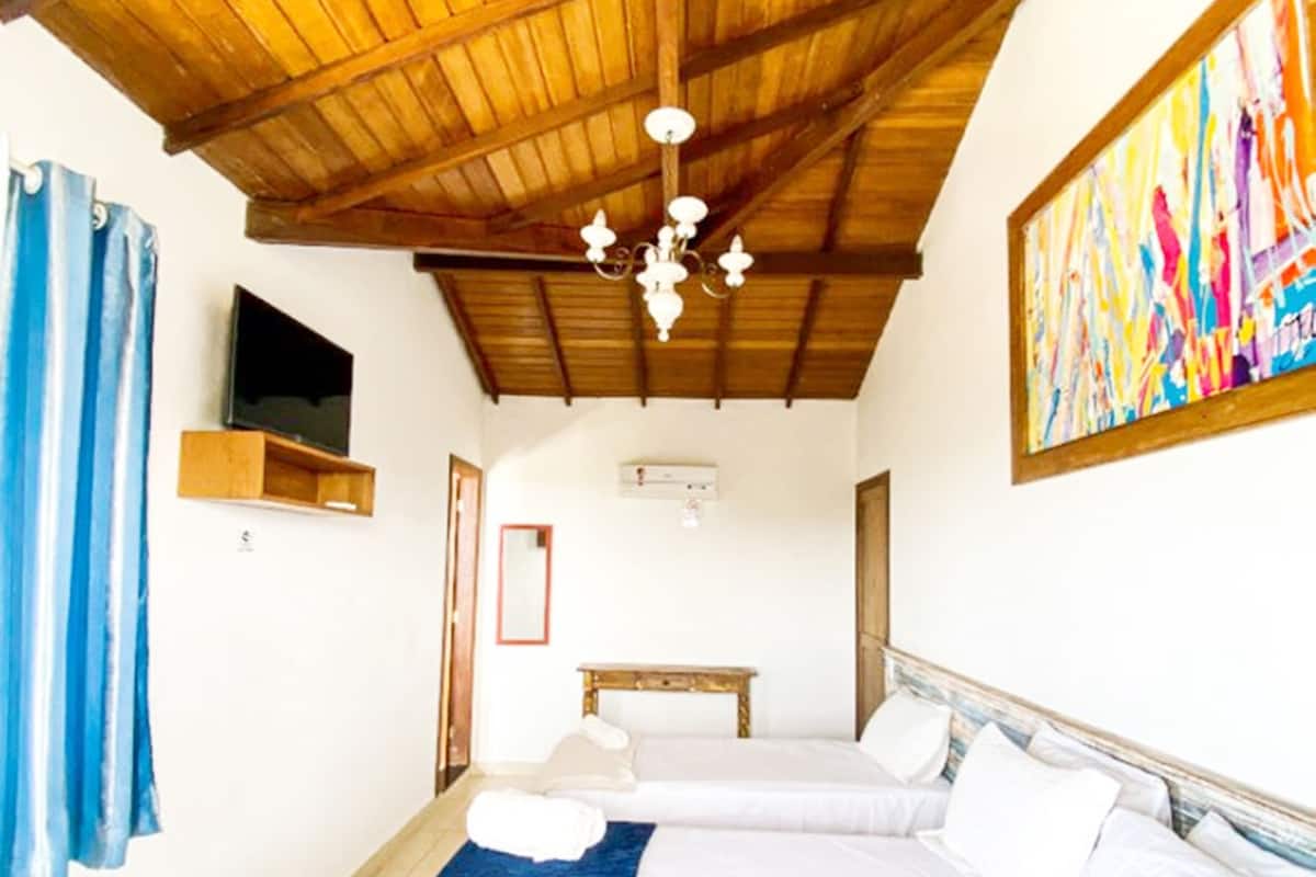 A bedroom with a wooden ceiling, a bed with white linens, a blue throw blanket, a painting on the wall, and a television mounted on the wall.