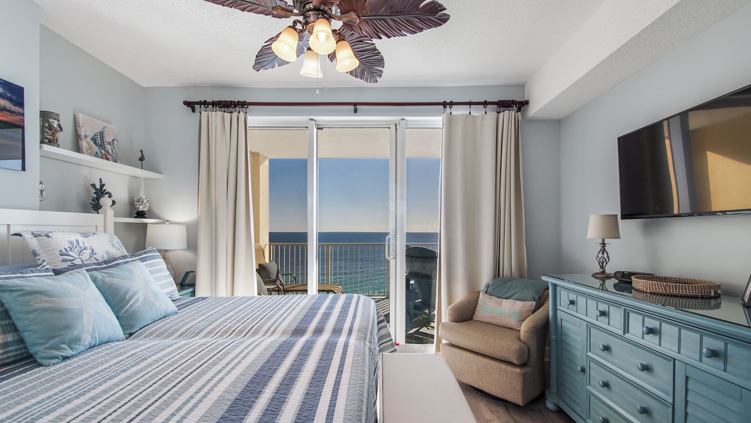 Een slaapkamer met een bed, een stoel, een dressoir en een televisie. Er is een balkon met uitzicht op de oceaan.