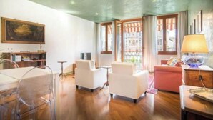 A spacious living room with a wooden floor, white sofas, a dining table with chairs, and a large window with curtains.