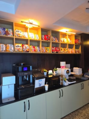 A snack bar with shelves of packaged snacks, a coffee machine, an ice dispenser, and a small plant. - shincheon noble stay hotel (Seoul)