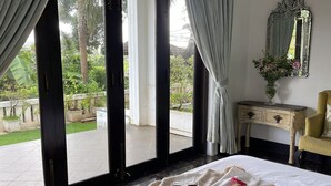 A room with a bed, a bedside table with a vase of flowers, and a view of a garden through large windows. - Villa One - Pvt Pool Outdoor Deck North Goa (Nerul)