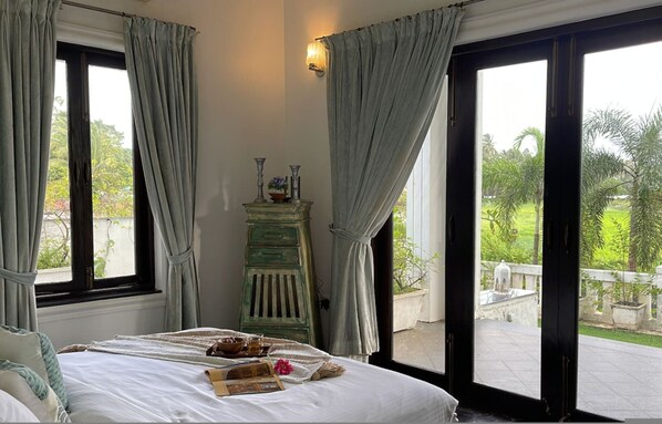 A bedroom with a bed, a nightstand, a lamp, and a view of a garden through a sliding door. - Villa One - Pvt Pool Outdoor Deck North Goa (Nerul)