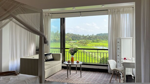A hotel room with a large window, a bed, a sofa, a small table with a vase, and a desk with a chair. - Villa One - Pvt Pool Outdoor Deck North Goa (Nerul)