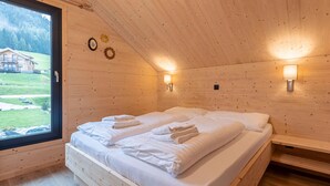 Ett sovrum i en stuga med träpanel, en stor säng, två sänglampor och ett fönster med utsikt över ett hus och berg.