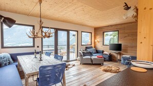 Un soggiorno moderno con soffitto in legno, zona pranzo con tavolo e sedie, divano, televisore e un'ampia finestra con vista sulle montagne.