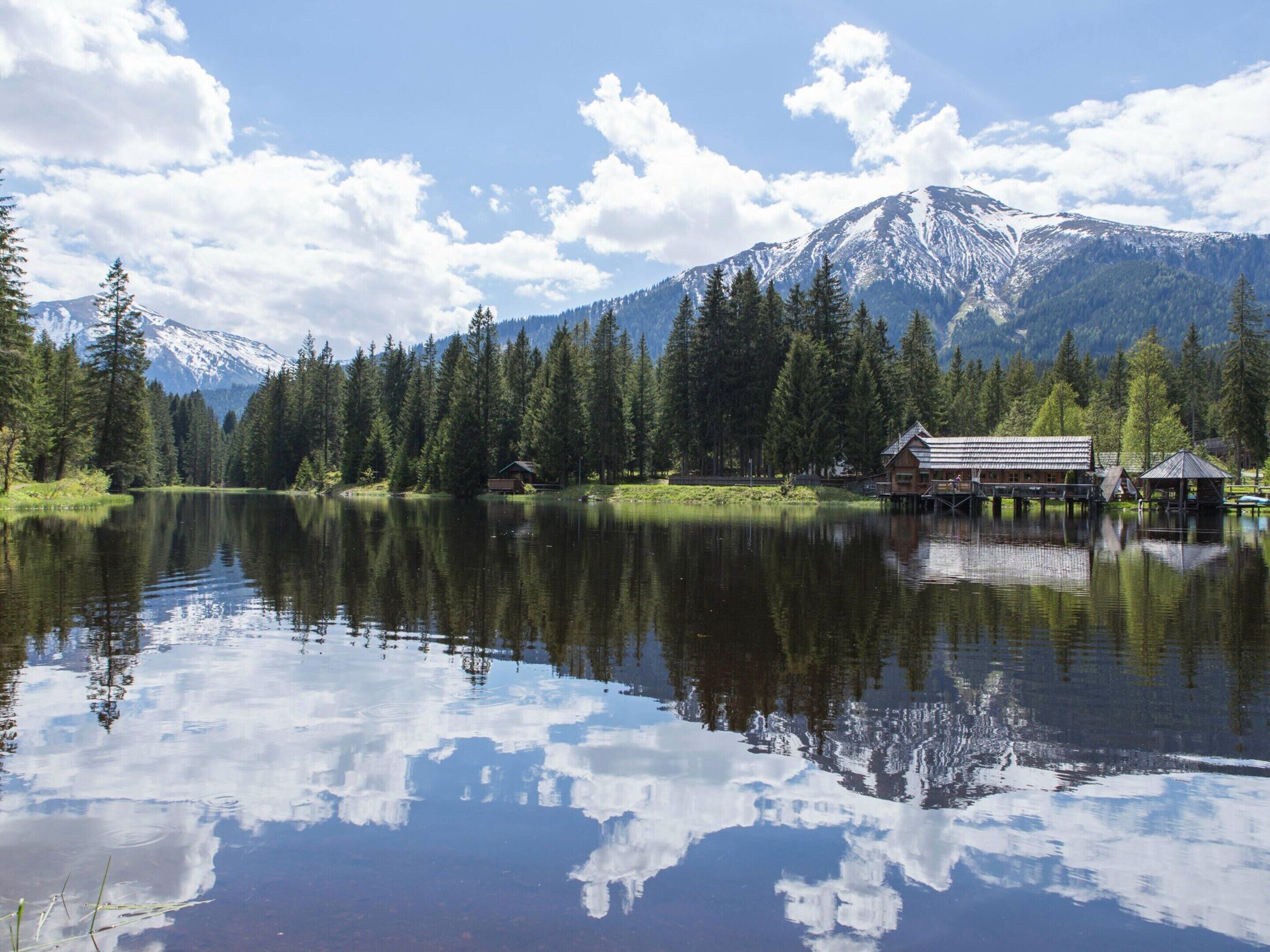 A tranquil lake with reflections of trees and mountains, surrounded by a forest and a cabin with a dock.
