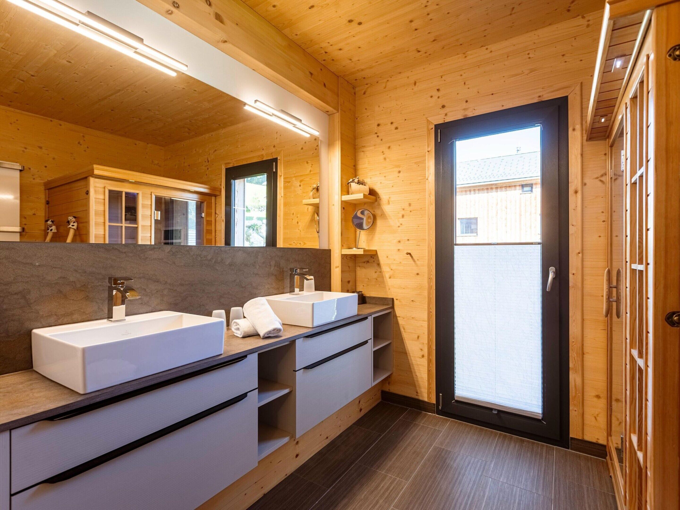 A modern bathroom with wooden walls, a large mirror, two sinks, and a door leading outside.