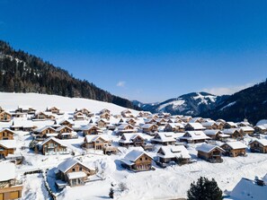 Ein verschneites Dorf mit Holzhütten, blauem Himmel und Bergen im Hintergrund.