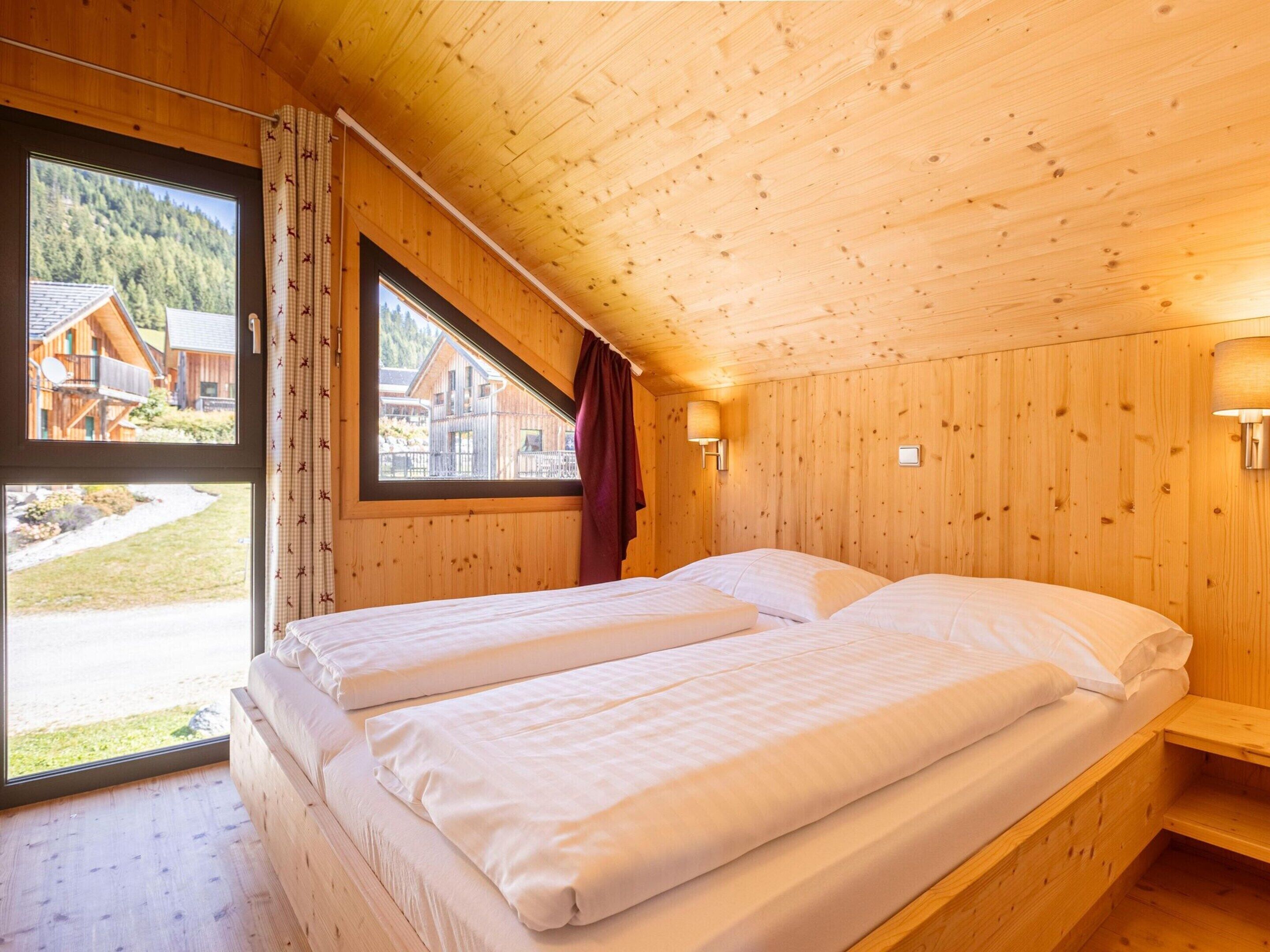 A wooden cabin bedroom with two beds, large windows, and a view of a wooden house and forest.