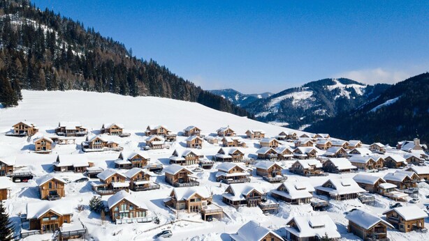 Ein schneebedecktes Dorf mit Holzhäusern, einer Skipiste und Bergen im Hintergrund.