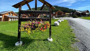 Un panneau en bois sur lequel sont inscrits les mots «ALMDRIFT» et «HOHNTALER», entouré de fleurs et placé sur un fond composé de maisons et d’un ciel dégagé.