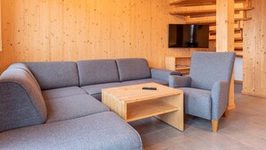 Un salon moderne avec un canapé d’angle gris, une table basse en bois et un fauteuil gris. Un téléviseur à écran plat est fixé au mur, au-dessus du canapé.