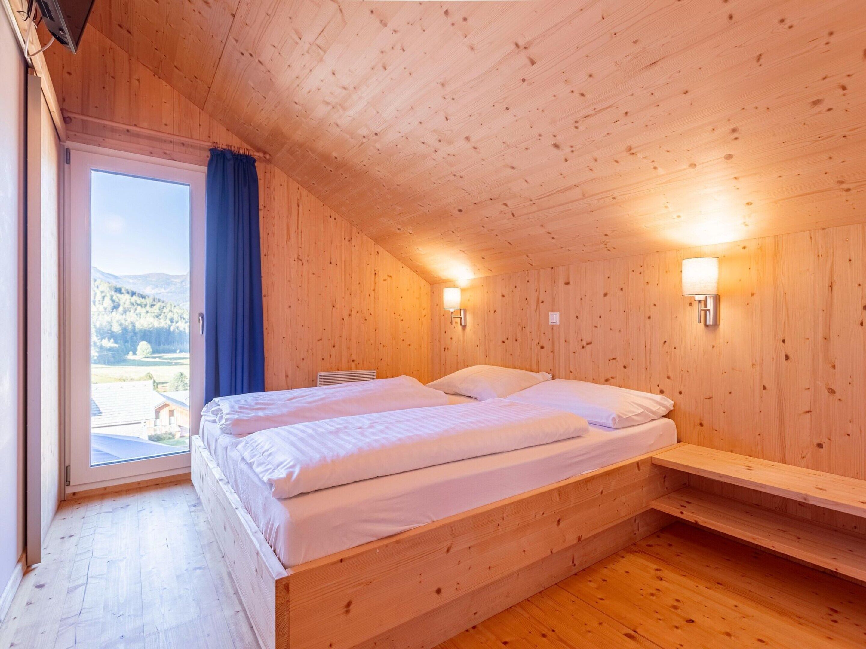 A wooden room with a bed, a window with a view of the outdoors, and two wall-mounted lights.