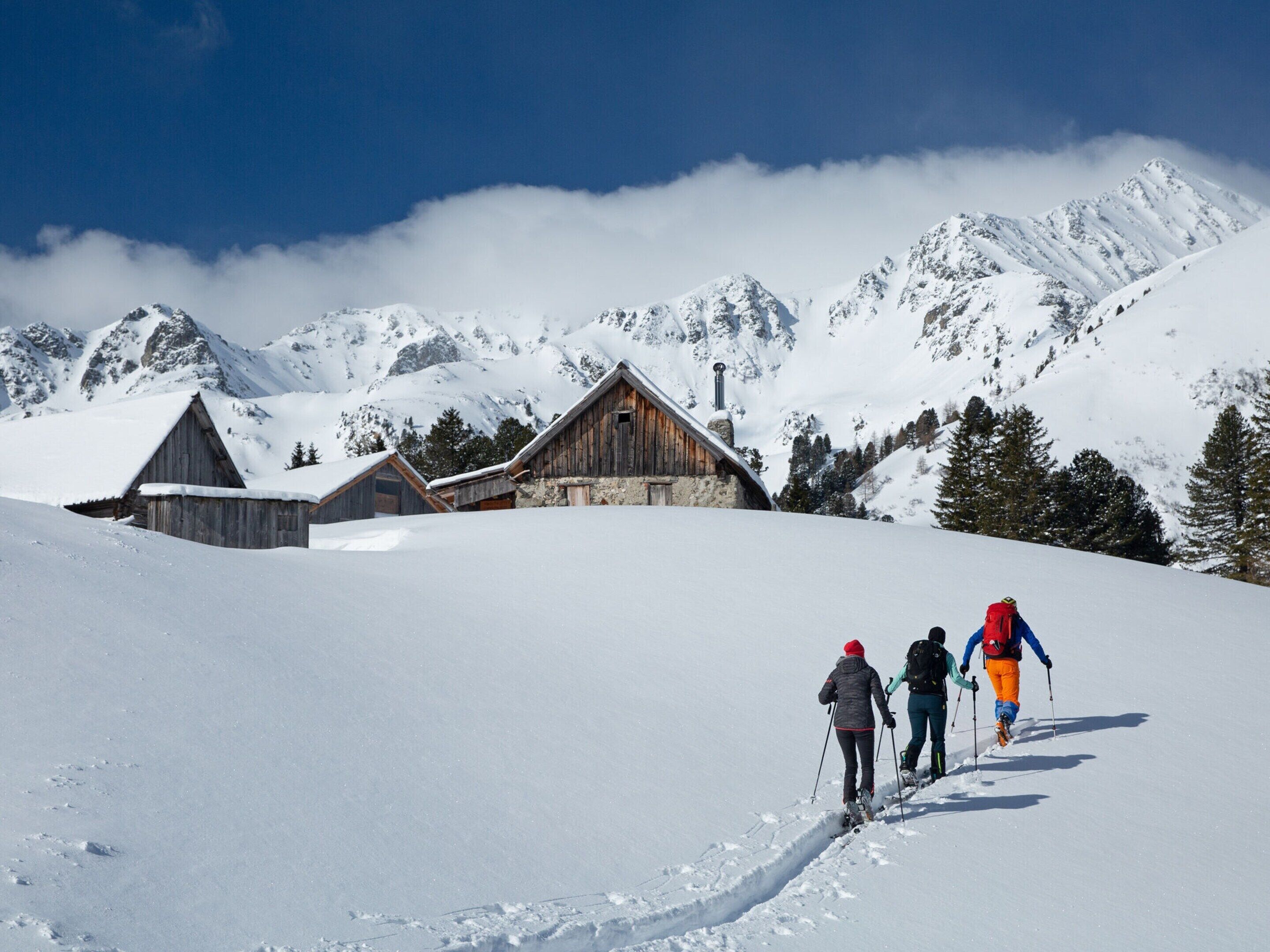 Une cabane en bois, construite sur des fondations en pierre, entourée de montagnes et d’arbres couverts de neige.