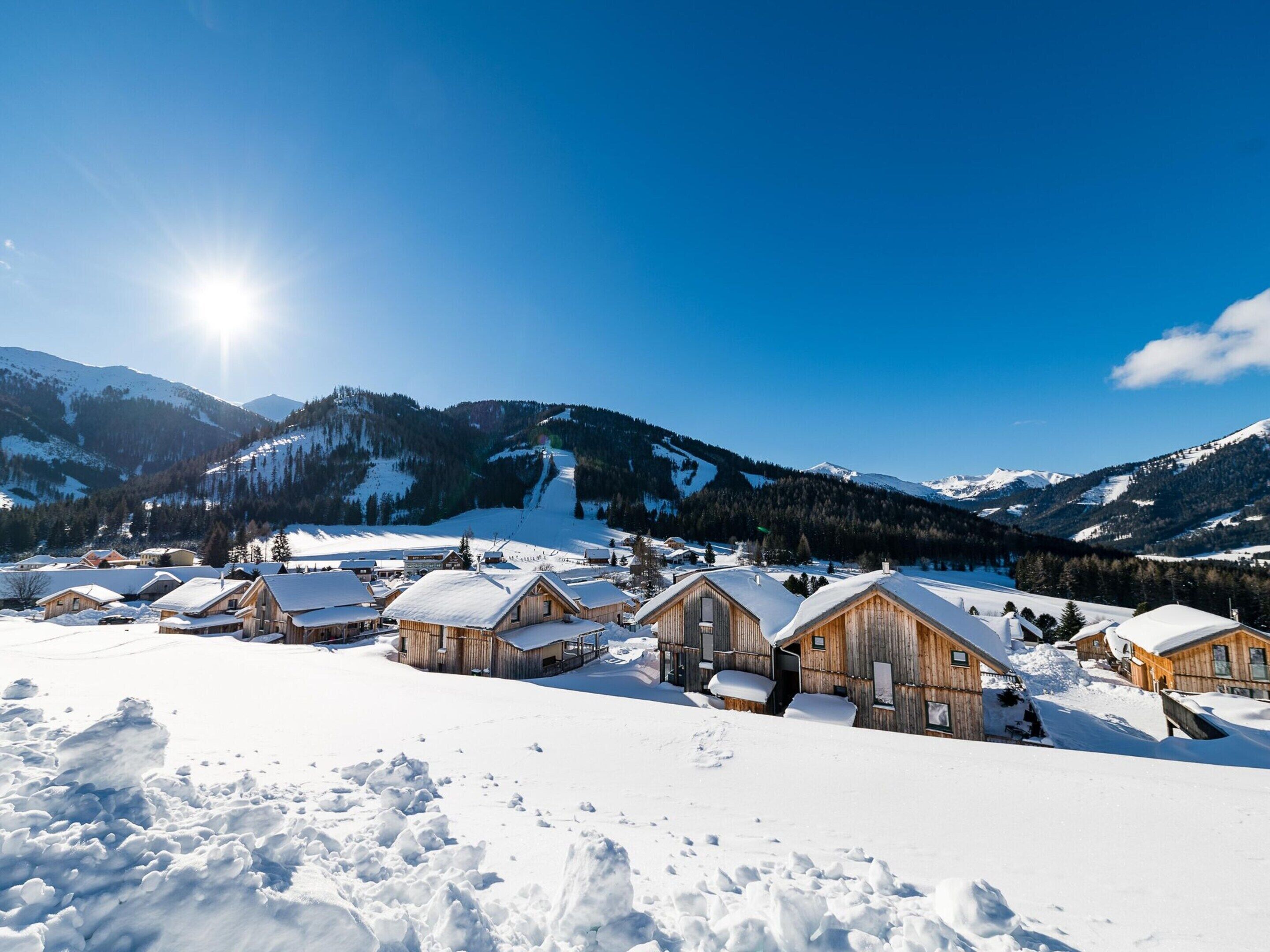 A snow-covered village with wooden cabins, a ski slope in the background, and mountains under a clear sky.