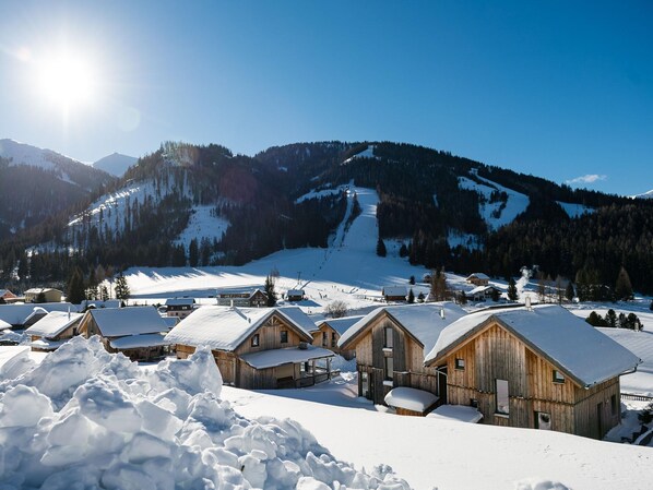 Un pueblo cubierto de nieve con cabañas de madera y una pista de esquí al fondo.