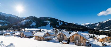 A snow-covered village with wooden cabins, a ski slope in the background, and mountains under a clear sky.