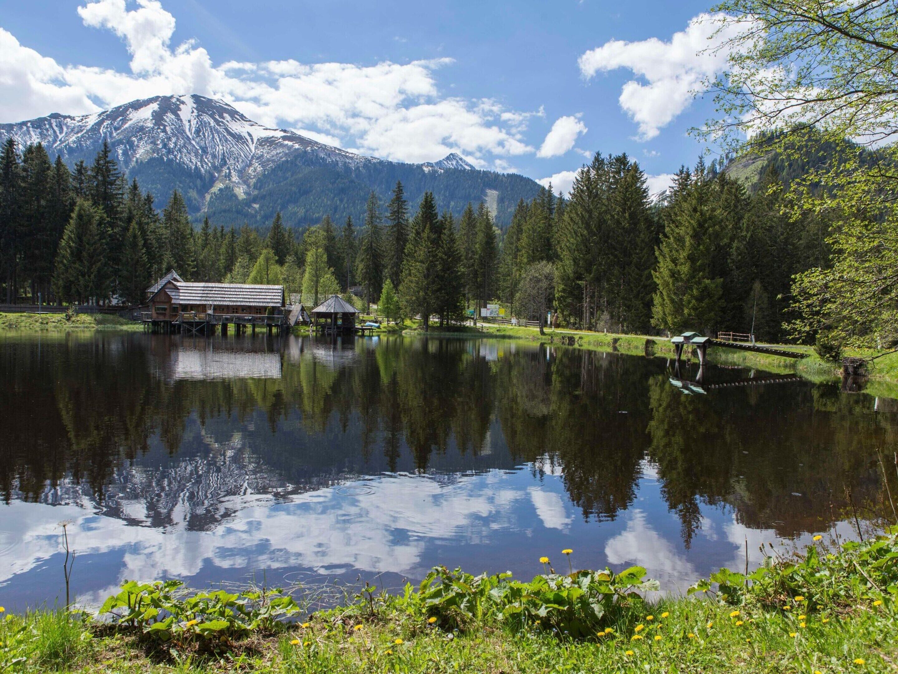 A cabin with a wooden deck over a lake, surrounded by dense forest and a snow-capped mountain in the background.