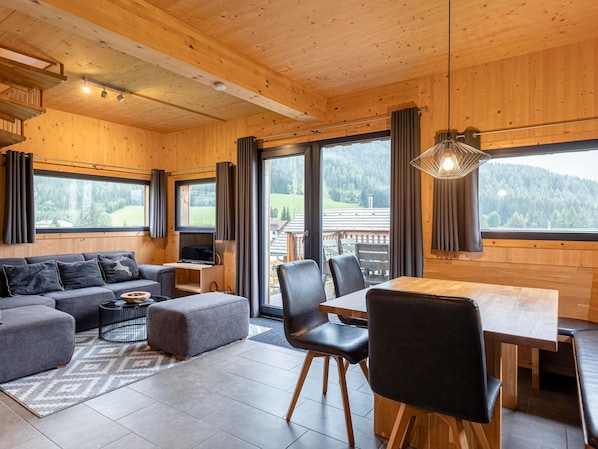 Interior moderno de una cabaña de troncos con sofá, reposapiés, mesa de comedor, sillas y televisor.