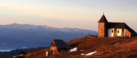 A church with a bell tower on a hillside with snow patches and a mountain range in the background.
