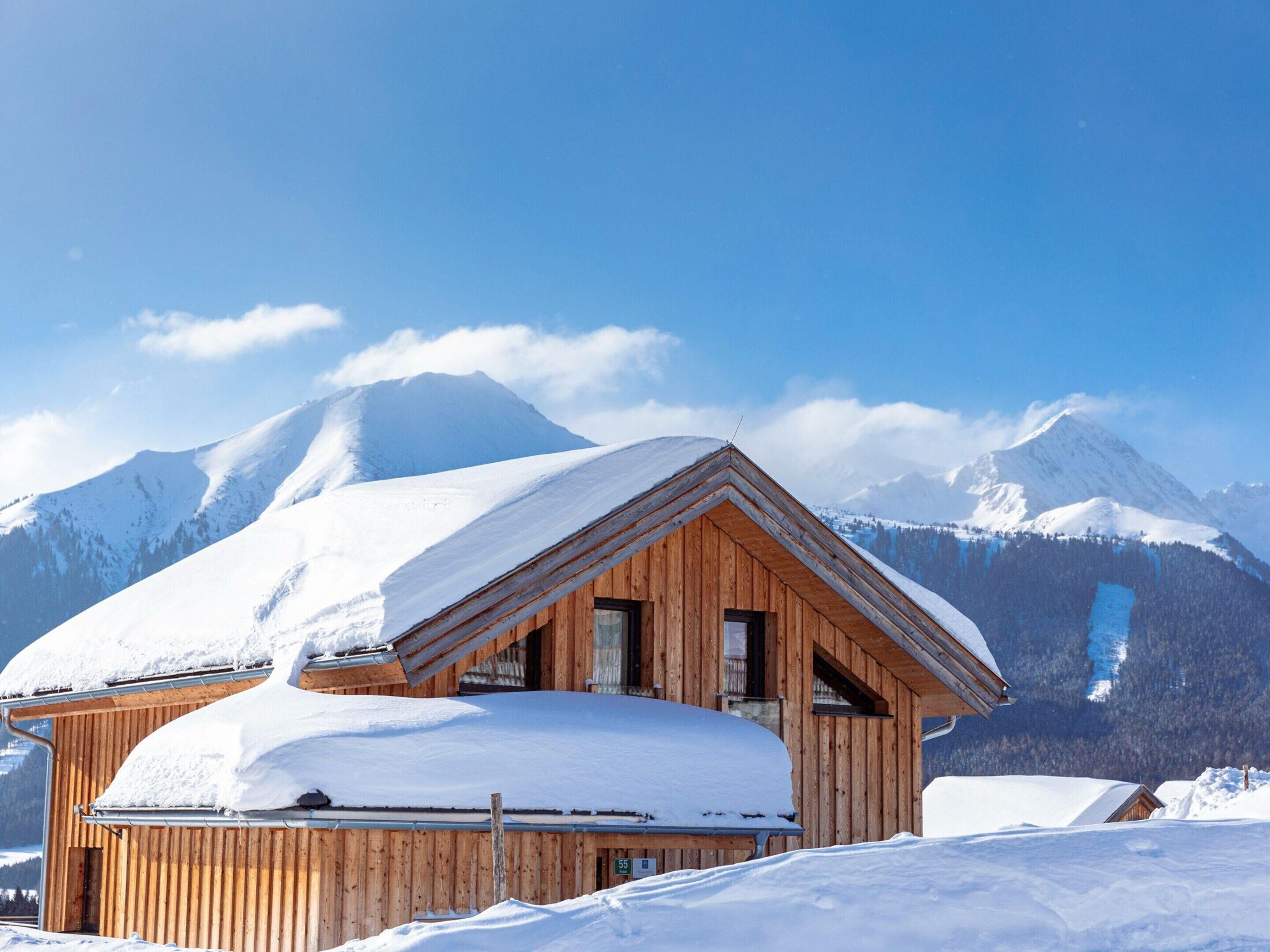 A wooden chalet with a sloped roof covered in snow, set against a backdrop of snow-capped mountains and a clear blue sky.