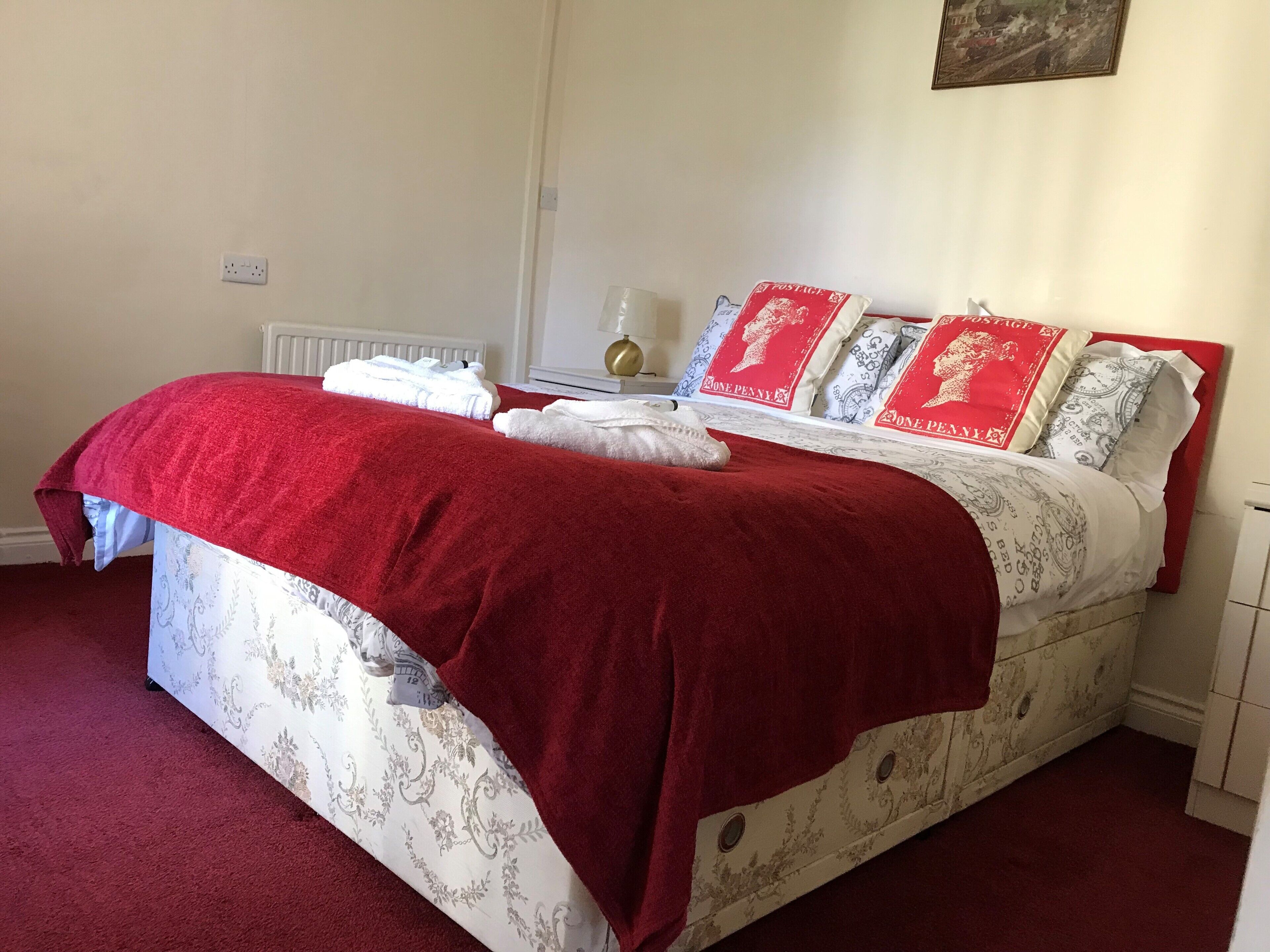 Een net opgemaakt bed met een rode deken en witte lakens, een radiator en een aan de muur hangend schilderij.
