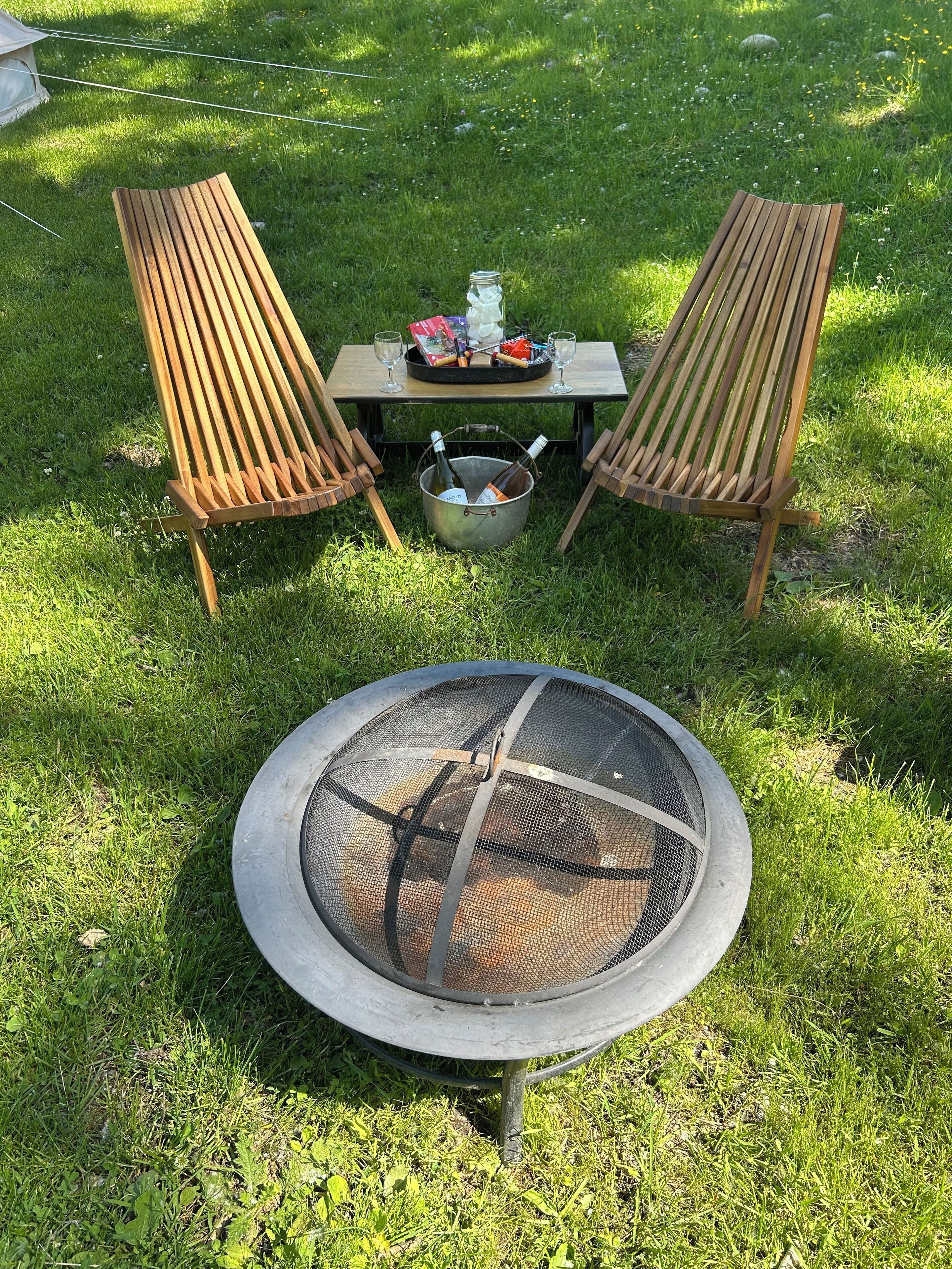 A grassy outdoor area with two wooden chairs, a table with a tray, and a metal fire pit.