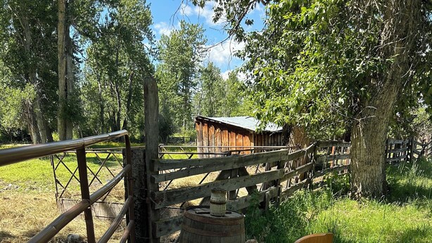 Un banco de madera con cojines y un tapete tejido, una cerca rústica de madera y una estructura de madera en un área boscosa y verde.