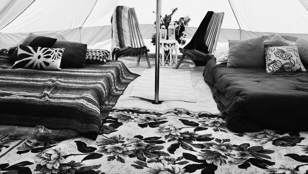 Une tente avec un poteau central et des parois en tissu, aménagée avec deux lits et une chaise, et un tapis à motifs sur le sol.