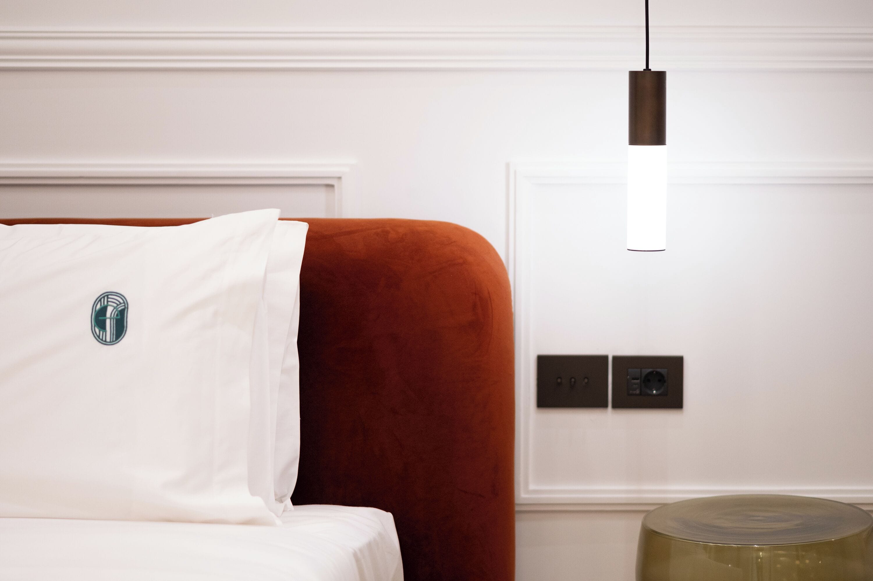 Ett hotellrum med en säng, en röd, klädd sänggavel, en vit kudde, en hängande lampa och ett litet, runt bord.