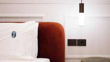 Ett hotellrum med en säng, en röd, klädd sänggavel, en vit kudde, en hängande lampa och ett litet, runt bord.