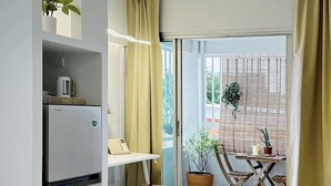 A compact kitchen with a built-in refrigerator, a small table, and a bench. A sliding glass door leads to a balcony with a potted plant and a view of the outside. - UDMS On nut 10 (Bangkok)