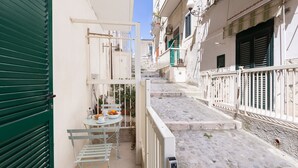 Un vicolo stretto con un tavolo e sedie su un balcone, una porta con persiana verde e una scala che conduce a un altro edificio.