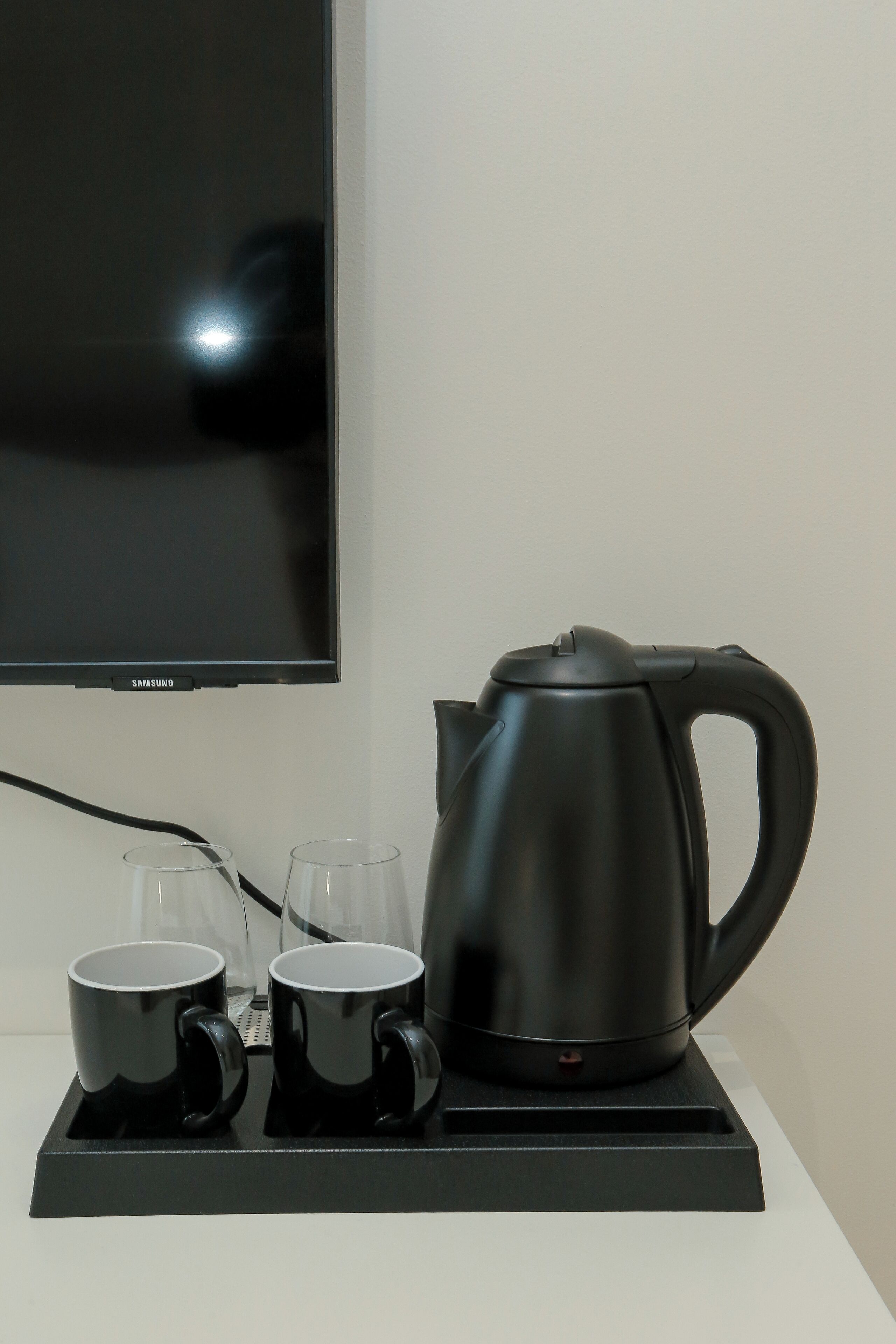 Une bouilloire noire est posée sur un plateau, à côté de deux tasses noires, devant un téléviseur Samsung.